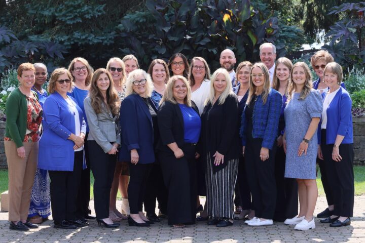 A group of 21 people, dressed in professional attire, poses outdoors on a sunny day. They are standing on a stone walkway with lush greenery and trees in the background. The group includes a mix of men and women, most of whom are wearing shades of blue, black, and white, with a few in other colors. Everyone is smiling, and they appear to be enjoying the moment together in a casual and friendly atmosphere.