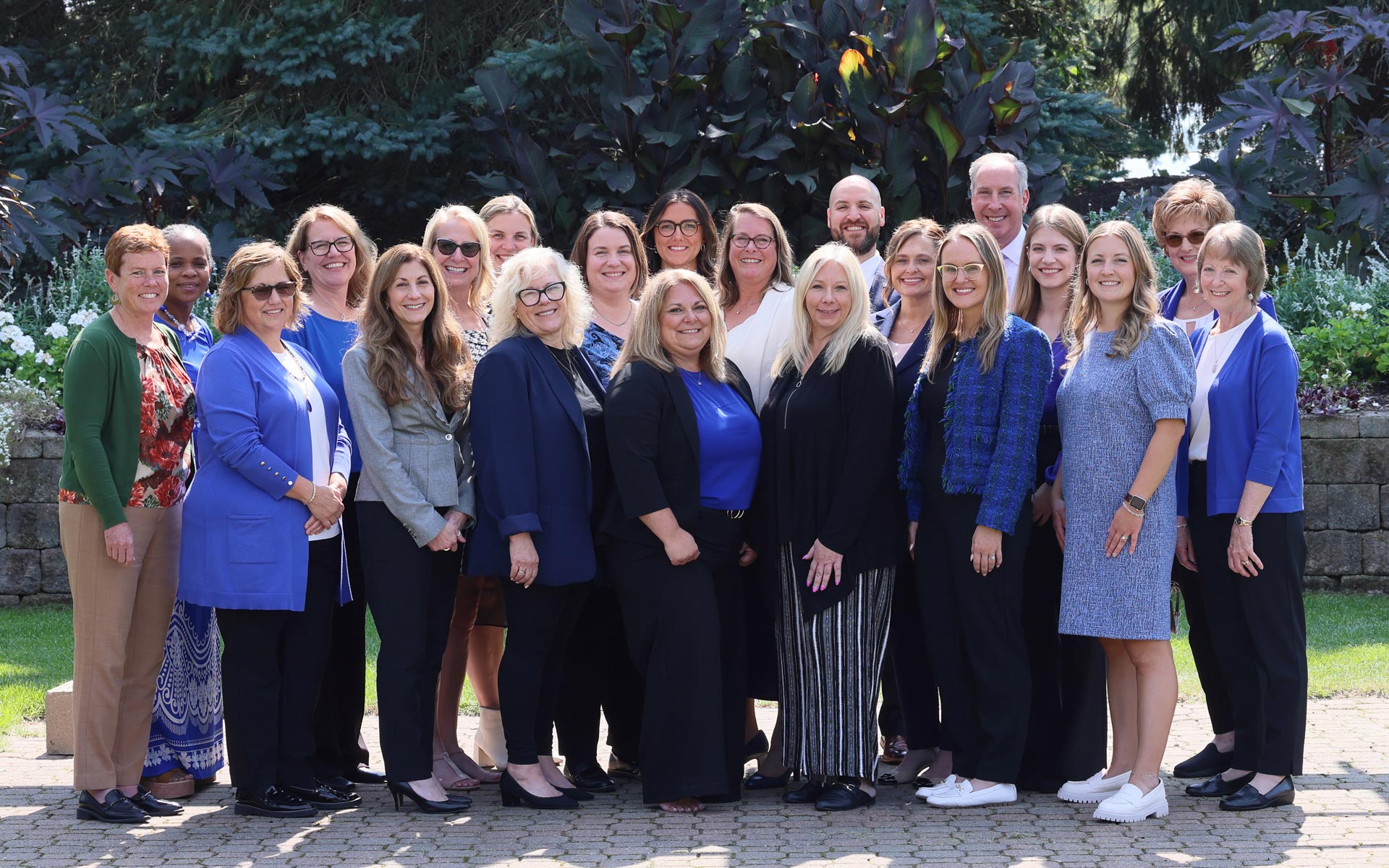 A group of 21 people, dressed in professional attire, poses outdoors on a sunny day. They are standing on a stone walkway with lush greenery and trees in the background. The group includes a mix of men and women, most of whom are wearing shades of blue, black, and white, with a few in other colors. Everyone is smiling, and they appear to be enjoying the moment together in a casual and friendly atmosphere.