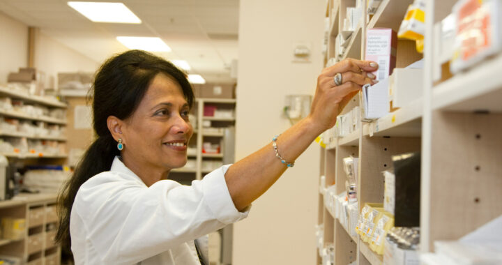 A pharmacist assisting a customer at a pharmacy, surrounded by shelves of medication and health products.