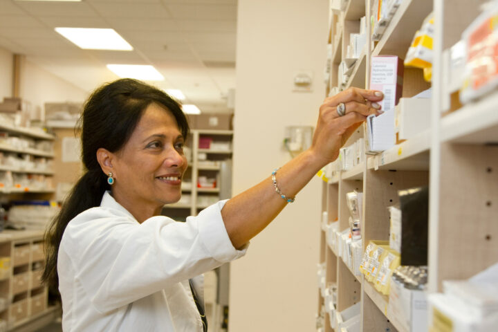 A pharmacist assisting a customer at a pharmacy, surrounded by shelves of medication and health products.