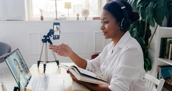 A woman sits at a wooden desk in a bright, modern room, wearing headphones and engaged in an online video call. She is looking at her laptop screen, which shows another person on the call. Her smartphone is mounted on a tripod, positioned to record or broadcast her side of the conversation. She holds an open notebook and gestures with her hand as she speaks. Behind her, a large plant and bookshelves with various items add a cozy touch to the workspace. Natural light fills the room through a large window.