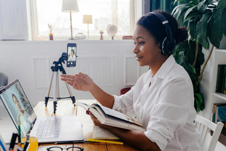 A woman sits at a wooden desk in a bright, modern room, wearing headphones and engaged in an online video call. She is looking at her laptop screen, which shows another person on the call. Her smartphone is mounted on a tripod, positioned to record or broadcast her side of the conversation. She holds an open notebook and gestures with her hand as she speaks. Behind her, a large plant and bookshelves with various items add a cozy touch to the workspace. Natural light fills the room through a large window.